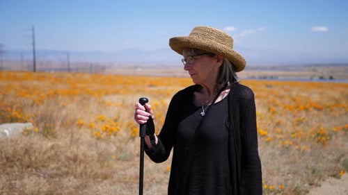 An older woman with a walking stick hiking in a field of flowers under a blue sky in slow motion.