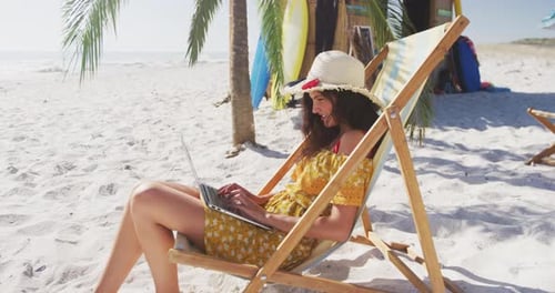 Kaukasische Frau sitzt auf einer Sonnenliege und benutzt seinen Laptop am Strand