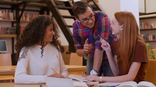 Two Teenage Girls Sitting at Library with Boy in Shirt and Glasses Standing Among Them Discussing