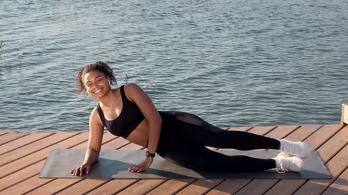 Young Happy Woman Lying on Pier and Practicing Side Leg Lift Exercise Exercising Outdoor Near Water