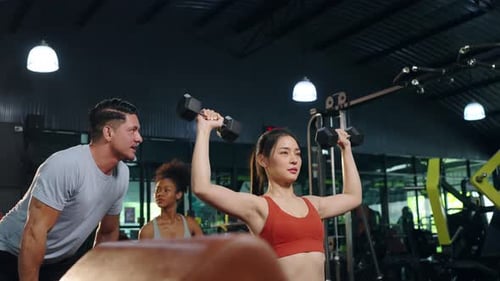 4K Asian woman do weight training workout lifting dumbbell with male sport trainer at gym.