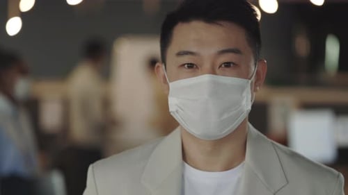 Young Asian Handsome Businessman in Medical Mask Looking at the Camera Standing in Modern Office