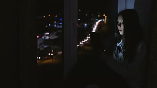 Glückliche Frau nutzt Tablet für Video-Chats und sitzt nachts im Dunkeln auf der Fensterbank