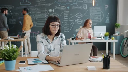 Female Entrepreneur Using Laptop in Shared Office While Coworkers Busy in Background