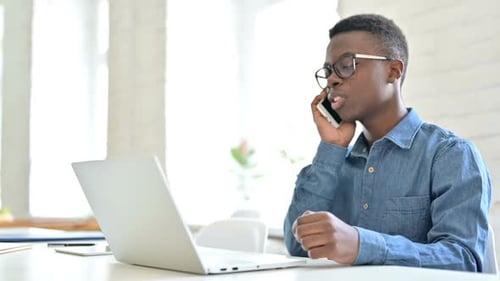 Young African Man with Laptop Talking on Smartphone in Office