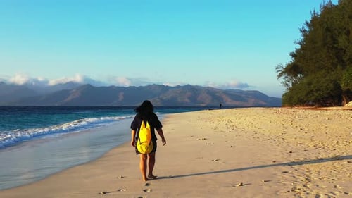 Girl walking alone on quiet beach with white sand alongside sea waves at sunrise with mountains of f