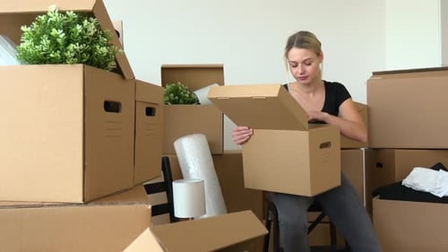 A Moving Woman Sits on a Chair and Puts Things Into a Cardboard Box in an Empty Apartment