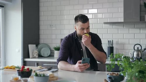 Bearded Man Using Phone and Eating Apple in Kitchen