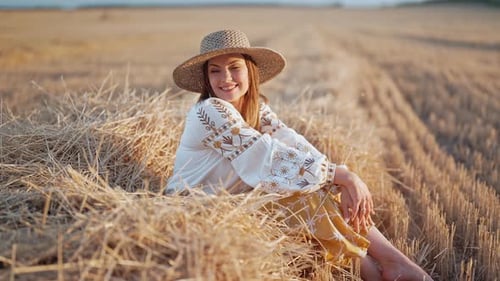 Beautiful Ukrainian Woman in Straw Hat and Vyshyvanka Blouse Posing Lying on Hay in Countryside at