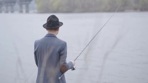 Junger Fischer in einer Jacke und einer Mütze mit Krempe am frühen Morgen beim Angeln im Fluss