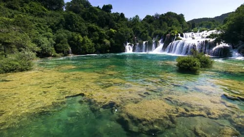 Tilt up from colorful water to wide waterfalls and forest at Krka