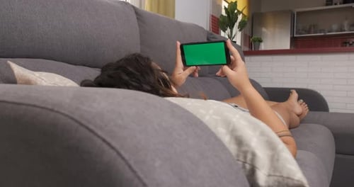 woman on the living room couch watching something on her cell phone, green screen on smart phone