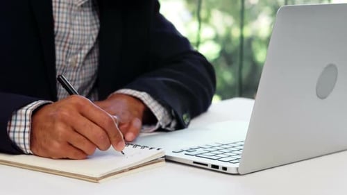 Man Writing in Notebook Beside Laptop at Table