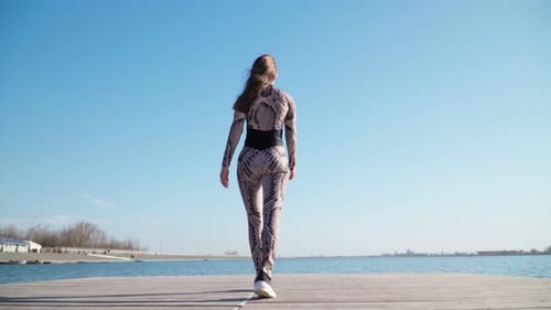 Young slim girl in sportswear is on the pier along the lake. Slow motion.