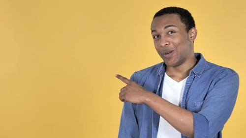 Young African Man Pointing with Finger on Side, Yellow Background