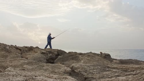 Viejo pescador de pie en las rocas junto al mar y pescando contra la puesta de sol
