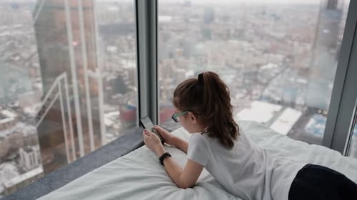 Cute Teenage Girl Surfing the Internet on a Mobile Phone While Lying on the Bed Near the Window City