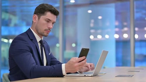 Businessman Working on Laptop and Holding Smartphone in Office