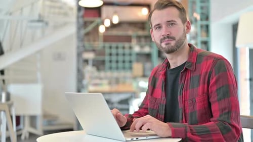 Young Man with Laptop Smiling at Camera in Cafe