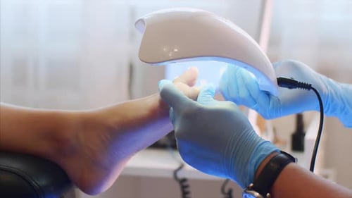 Le maître en pédicure en gants sèche la gomme-laque sur les orteils avec une lampe UV spéciale