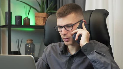 Young man is talking on smartphone in front of laptop computer in modern office