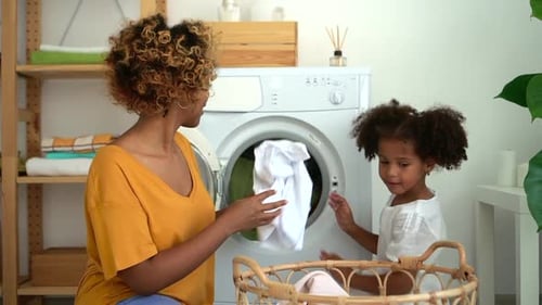 Young Mother and Little Daughter Putting Clothes in Washing Machine in Laundry Room at Home Spbd