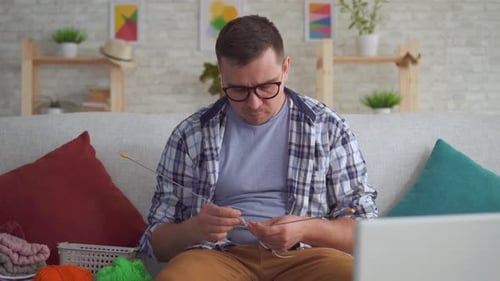 Close Up Young Man with Spokes in His Hands Learns To Knit on a Training Video on a Laptop