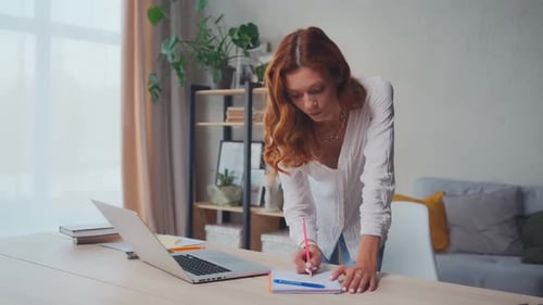 Woman Stands at Table Taking Notes from Laptop