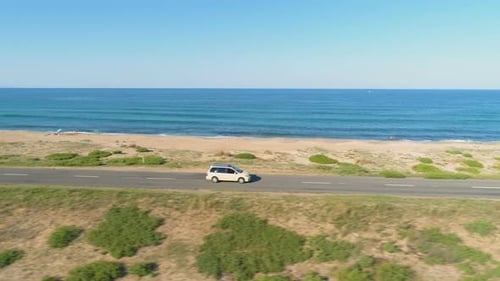 White Car Speeding on Straight Asphalt Coastal Road Against Blue Sea and Sand Beach