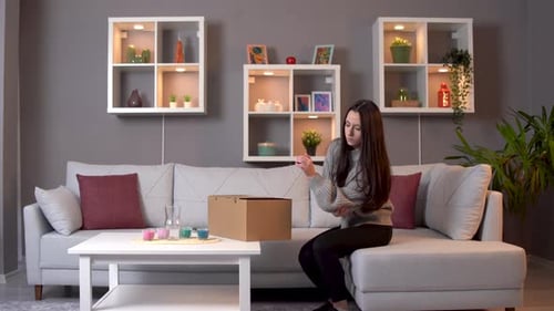 Woman Carries Box, Sits on Couch, Examines Hand