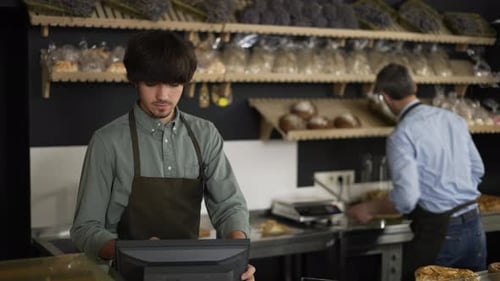 Young Cashier in Apron Typing on Touch Screen Handsome Bakery Worker Slicing Bread in the Background