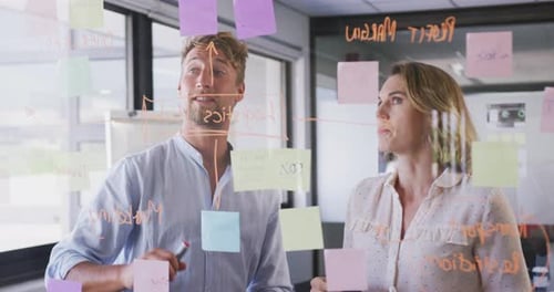 Professional businesswoman and businessman discussing over memo notes on glassboard in modern office