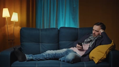 Man Lying on the Couch in a Cozy Room and Using Smartphone for Surfing Internet or Chatting in