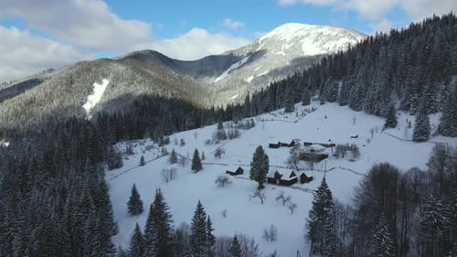 Aerial winter landscape with small rural houses between snow covered forest in cold mountains.