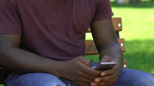 Concentrated Afro-American Man Sitting on Bench in Park, Using His Smartphone