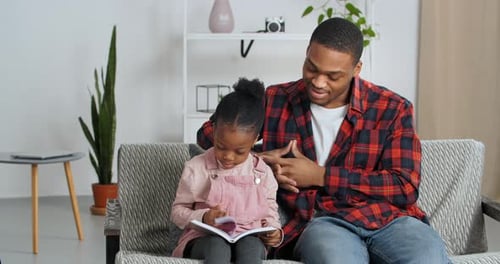 Afro American Father Sits with Beloved Daughter on Couch in Interior of Modern Living Room Kid Holds