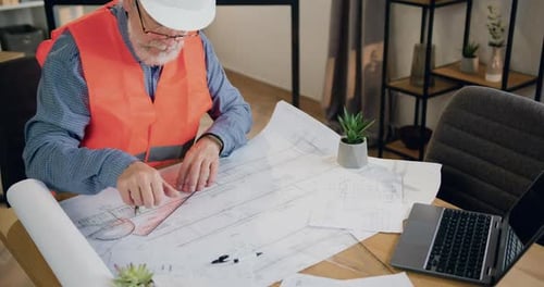 Senior Bearded Architect in Vest and Helmet which Drawing on Blueprint in Modern Design Office