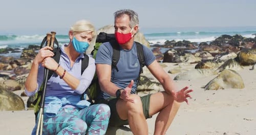 Senior hiker couple wearing face masks with backpacks and hiking poles talking sitting on rock