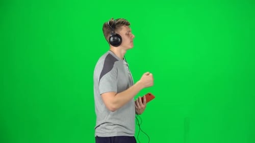 Man in Big Headphones Goes and Dances with Smartphone on Green Screen at Studio. Side View. Slow