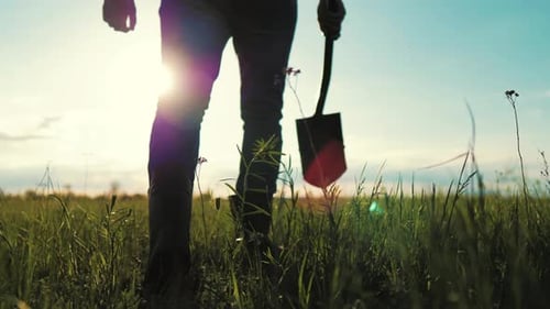 Farmer Walking with Shovel Across Grassy Green Field