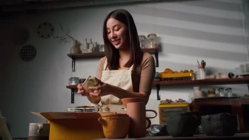 A young Asian woman ceramist is packing ceramic mugs in a parcel box for people who buy online.