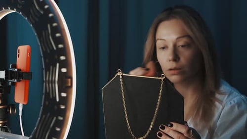 Blonde Female Fashion Blogger Applies Makeup in Front of Ring Light in Dark Room