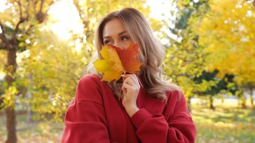 Woman in Red Dress Hides Face with Yellow Leaves in Park