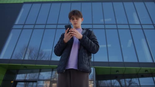 Medium Shot of Handsome Young Man Working on Smartphone Stands on the Street in Front of a Modern
