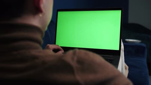 Over the Shoulder Shot of a Man Working Laptop on Sofa at Home with Green Screen, Chroma Key, Makes