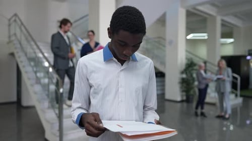 Young Intelligent African American Man Analyzing Paperwork Standing in Office Lobby with Colleagues
