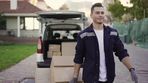 Junger gutaussehender lächelnder kaukasischer Zusteller mit Kartons auf dem Wagen, der mit geöffnetem LKW aus dem Van fährt