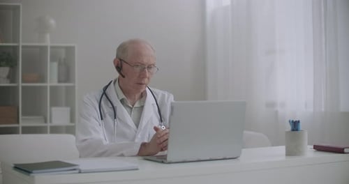 Älterer männlicher Arzt konsultiert Patienten online, sitzt im Büro der Klinik und kommuniziert durch