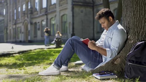 Gemischtrassiger junger Mann sitzt unter Baum, liest Interessantes Buch, Bücherwurm