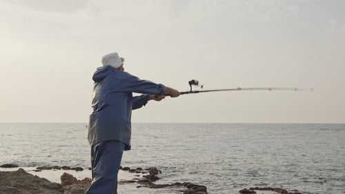 Old fisherman standing on sea side rocks and fishing against the sunset